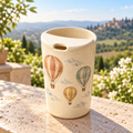 Digital concept of a speckled ceramic travel mug with terracotta, teal and ochre hot-air balloons on a stone ledge.